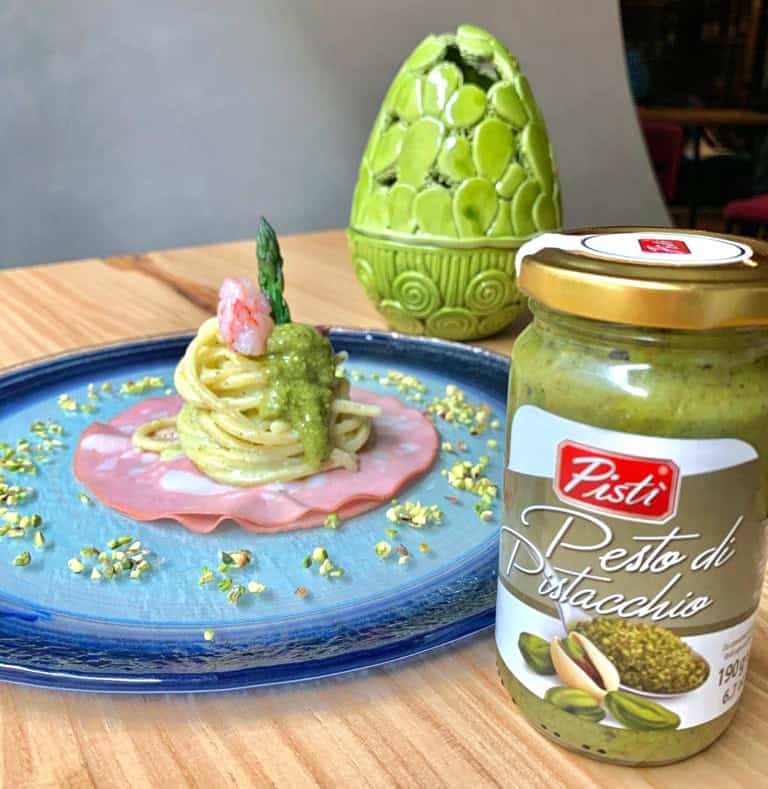 Pistì Sicilian Artisanal Pesto with Pasta Sicilian pistachio pesto with pasta on a blue plate, decorated with pistachios and pink ham.