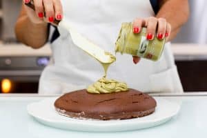 Person adding green pistachio cream to a chocolate cake, close-up shot.
