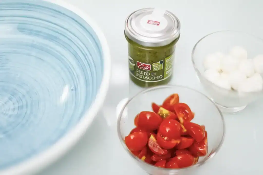 Pesto di Pistacchio e Pomodorini Pistachio pesto jar with cherry tomatoes and mozzarella balls on a white surface.