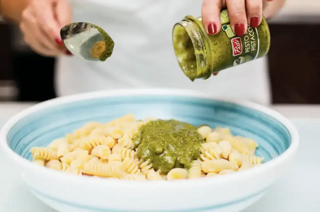 Pomodoro e pesto sulla pasta Adding green pesto to pasta in a blue bowl, close-up shot.