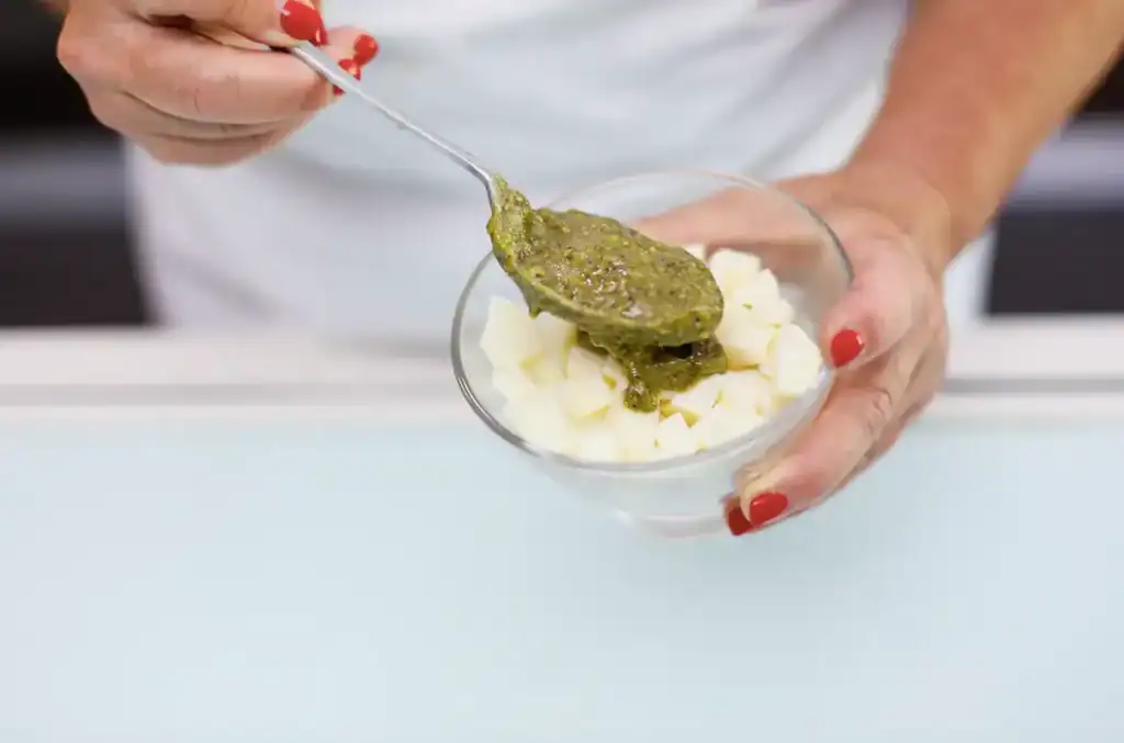 Pomodoro verde su purè di patate Person adding green pesto to mashed potatoes in a glass bowl.