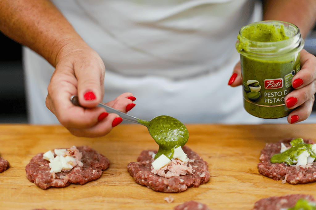 Pesto di Pistacchio Artisanal Woman spreading pistachio pesto on raw meat patties in a kitchen.
