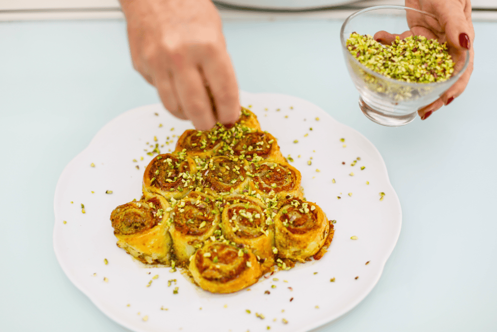 Pistì Sicilian Cannoli Rolls Hand sprinkling crushed pistachios on Sicilian cannoli rolls on a white plate.