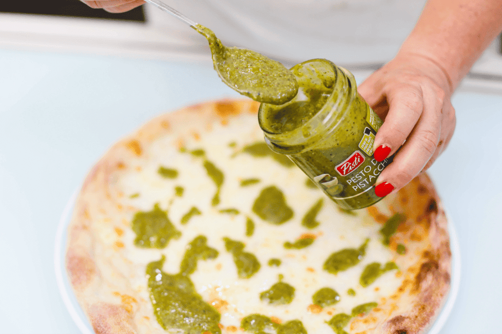 Pizza with green pesto being spread on it Pesto being spread on a pizza crust with a spoon.
