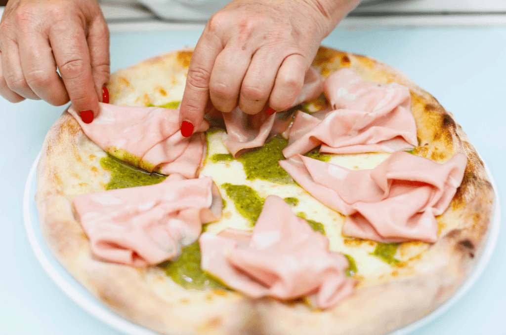 Pizza con prosciutto e pesto Hand placing prosciutto slices on a pizza with pesto sauce.