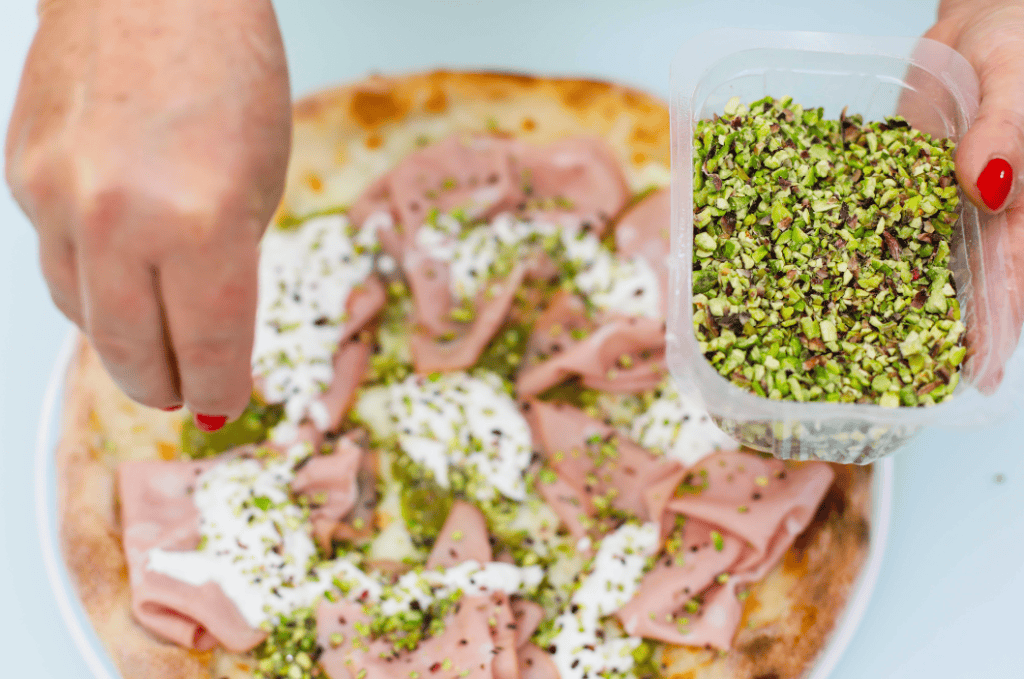Pizza with pistachios being garnished with chopped pistachios. Freshly baked pizza topped with slices of ham and sprinkled with chopped pistachios.