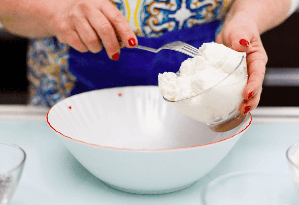 Filling a bowl with ricotta cheese Ricotta cheese being scooped into a bowl by a person with red nail polish.