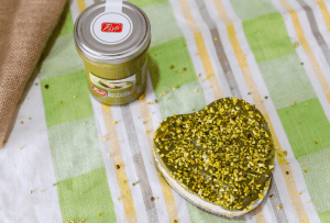Pistachio and tomato spread jar with pistachio-coated cheese on a checkered cloth.